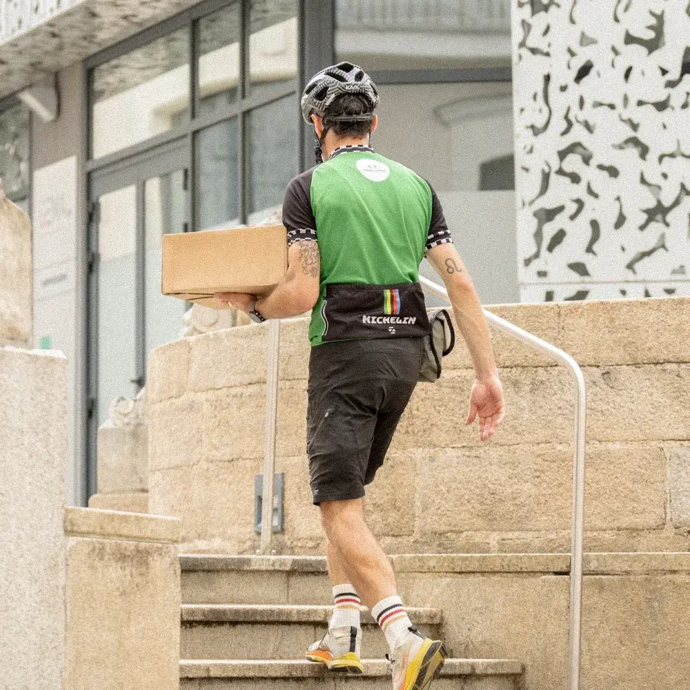 Un livreur, avec un maillot vert et un casque de vélo, portant un colis en carton se dirige vers son point livraison.